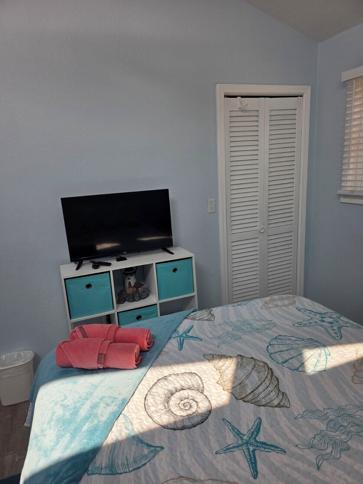 A bedroom with a bed, a TV on a white cabinet, a closet, and a window with blinds.