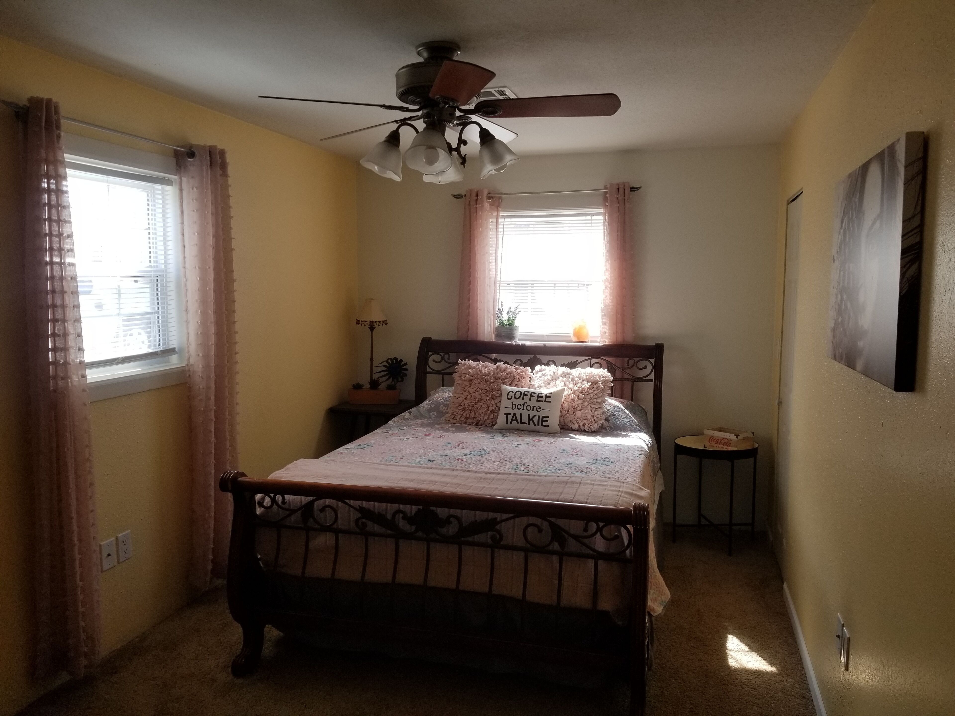 A bedroom with a bed, a ceiling fan, a nightstand, a lamp, and a window with curtains.