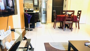 Een moderne woonkamer met een glazen salontafel, een rode eettafel met vier stoelen, een keuken met een koelkast en fornuis, en een flatscreen televisie aan de wand.