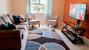 Een woonkamer met een bank, twee gestreepte stoelen, een salontafel, een televisie op een standaard en een groot vloerkleed.
