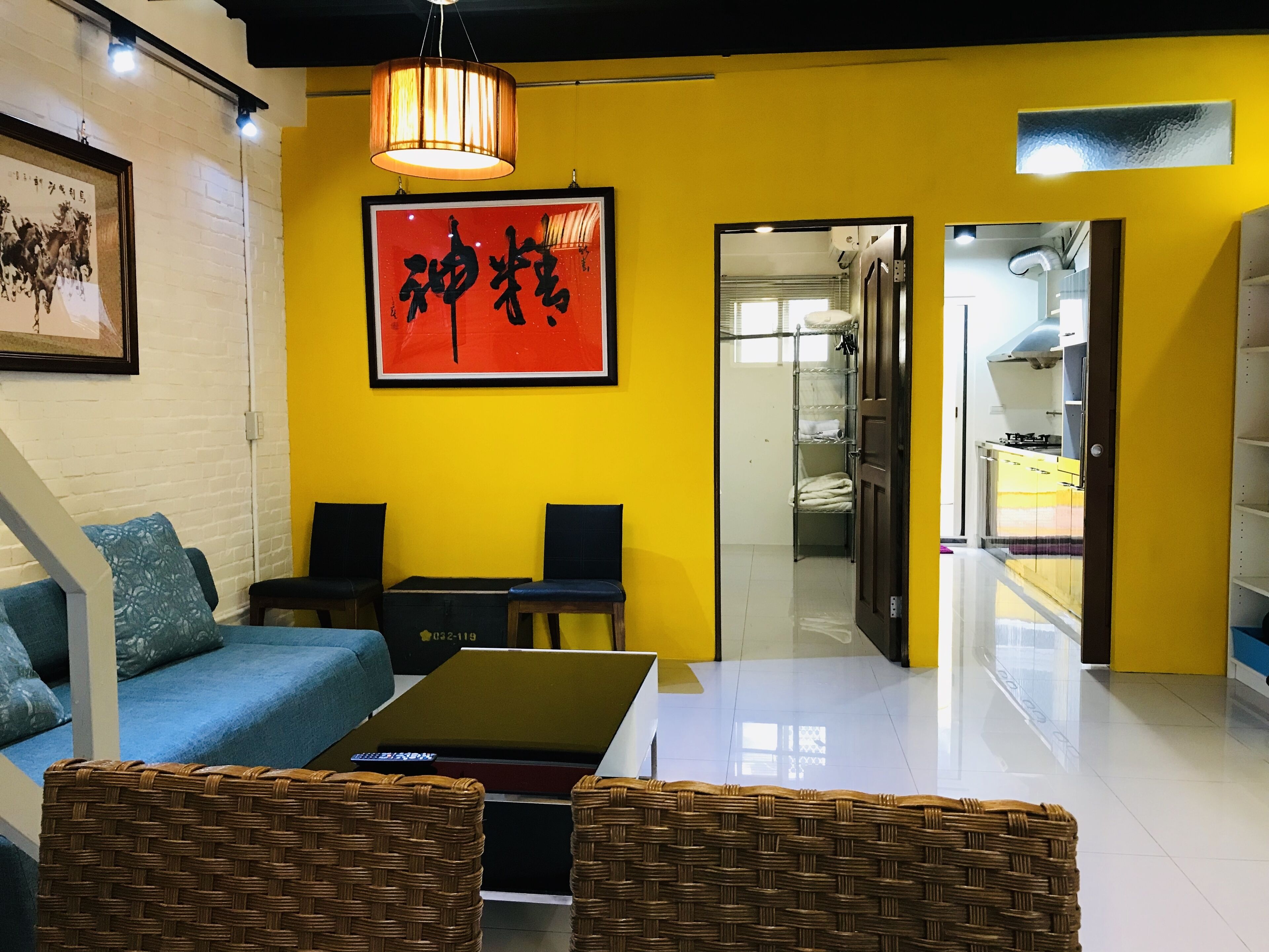A room with a yellow wall, a framed calligraphy piece, a sofa, a coffee table, and a view into a kitchen area.