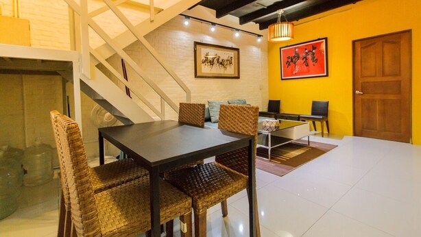 Un comedor con una mesa de madera oscura y sillas de mimbre, una escalera con barandillas blancas y una pared amarilla con un cuadro enmarcado.