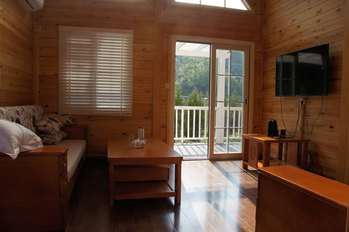 A wooden cabin room with a bed, a table, a TV, and a sliding glass door leading to a balcony.