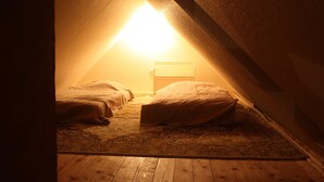Un pequeño dormitorio en el ático, tenuemente iluminado, con dos camas y una mesita de noche, que presenta techo inclinado y suelo de madera.
