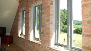 Una habitación con techo inclinado, paredes de ladrillo visto y ventanas grandes con marcos blancos.