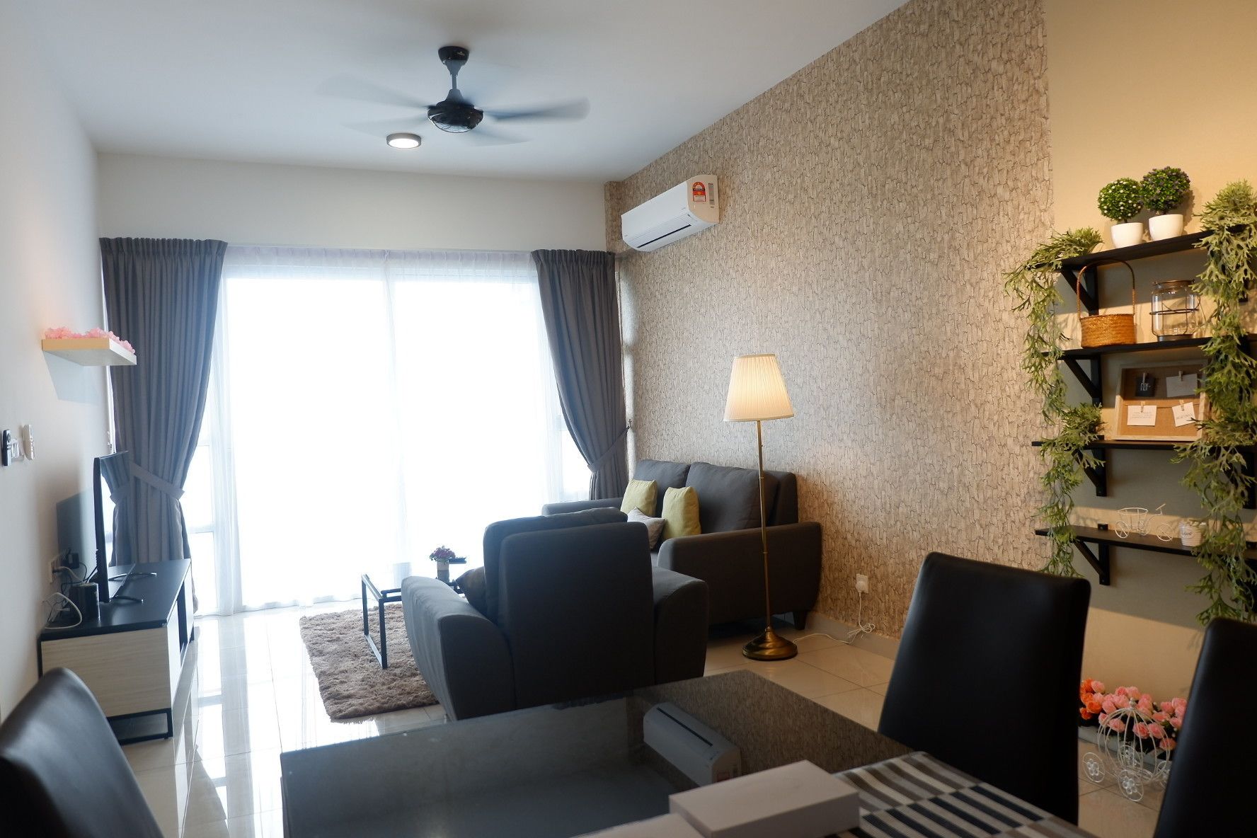 A modern living room with a sofa, armchairs, a coffee table, and a floor lamp. There is a ceiling fan, an air conditioning unit, and a window with curtains.