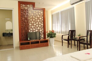Habitación de hotel con televisión sobre un soporte de madera, una planta en maceta, una mesa de comedor con sillas y un ventanal con persianas.