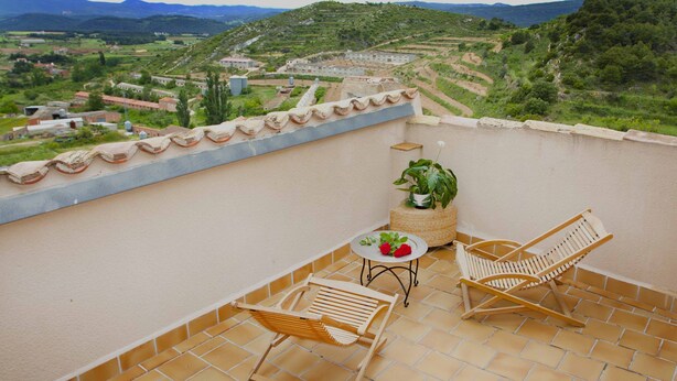 Eine Terrasse mit Blick auf eine hügelige Landschaft, zwei Holzstühle, ein kleiner Tisch mit einem Topfpflanzen und einer Blumenplatte.