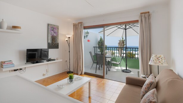 A modern living room with a TV, white shelves, a dining table, and a sofa. - Charming "La Florida" Apartment with Pool, Balcony, Sea Views & Wi-Fi (Tías)
