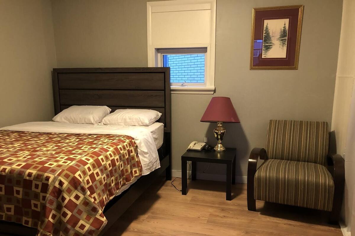 A bedroom with a bed, a nightstand with a lamp, a chair, and a framed picture on the wall.