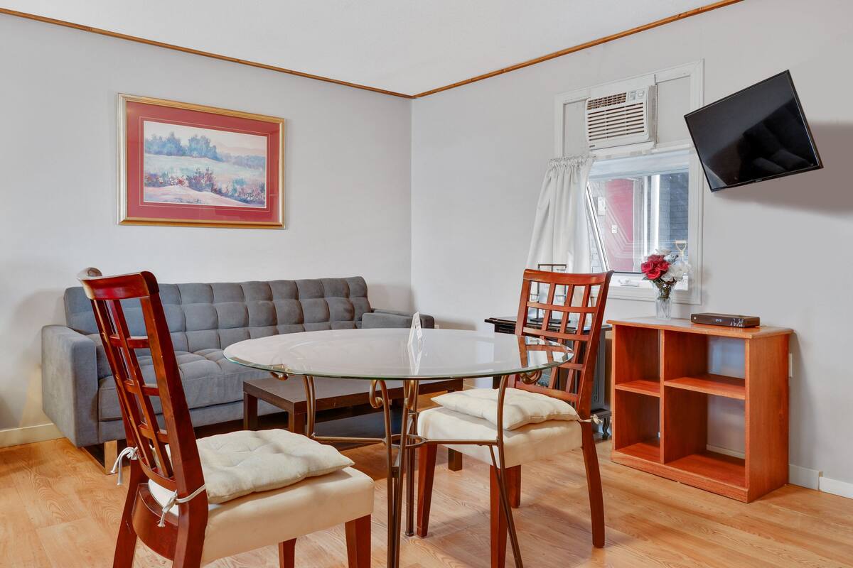 A dining area with a round glass table, four wooden chairs, a grey sofa, a wooden sideboard, and a mounted TV.