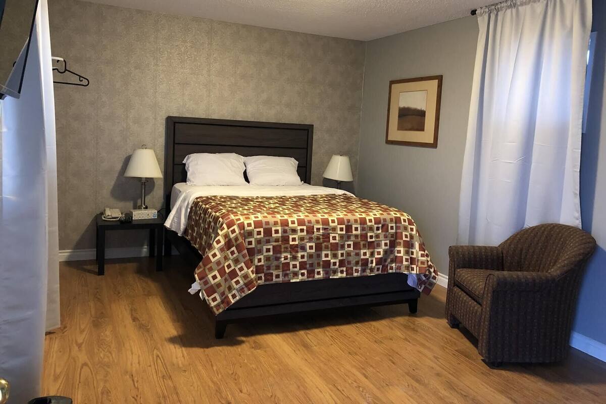A bedroom with a bed, two bedside lamps, a nightstand, a chair, and a framed picture on the wall.