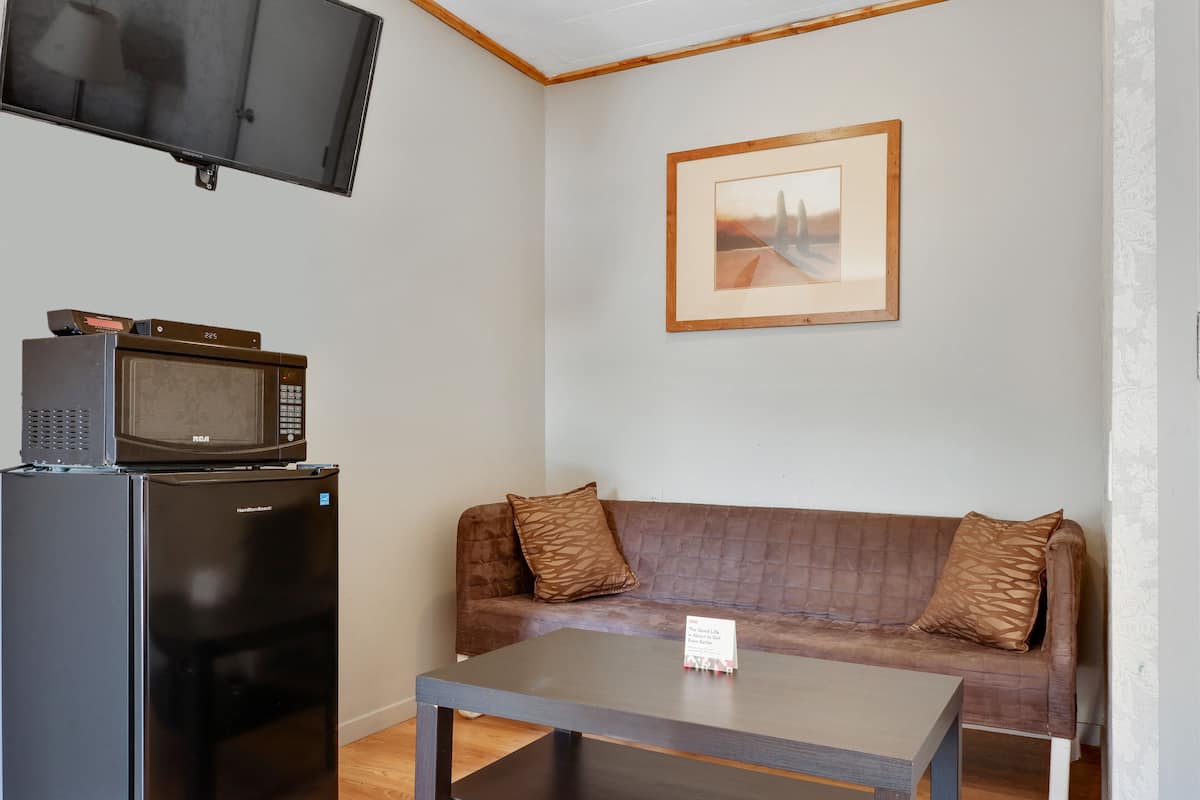 A compact living space with a flat-screen TV mounted on the wall, a microwave on a cabinet, a brown sofa with patterned cushions, and a small table with a sign.