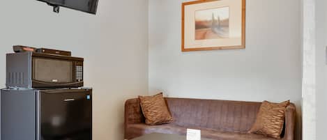 Un espacio habitable compacto con un televisor de pantalla plana montado en la pared, un microondas sobre un mueble, un sofá marrón con cojines estampados y una mesita con un letrero.