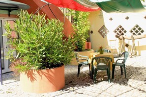 Une terrasse extérieure avec une table et des chaises, entourée de plantes en pot et agrémentée d’un parasol rouge.