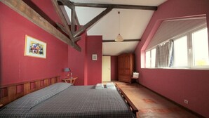Une chambre avec un grand lit, des poutres en bois, une fenêtre avec des rideaux et une porte donnant sur une autre pièce.