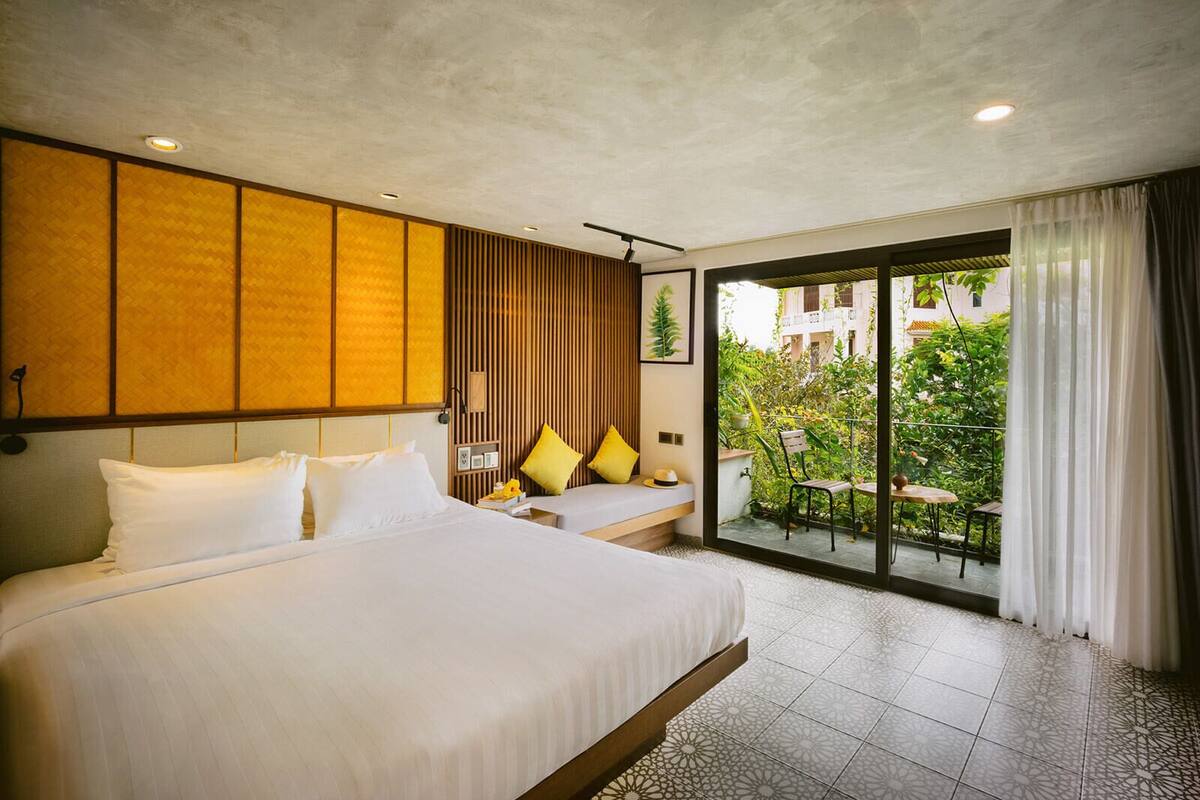 A hotel room with a large bed, a smaller bed, a balcony with a table and chairs, and a view of greenery outside.