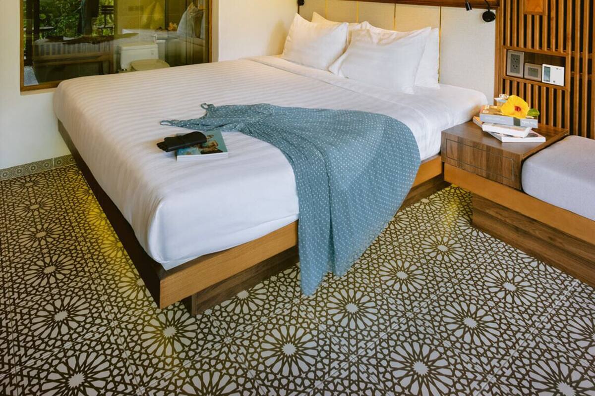 A modern hotel room with a large bed, a bedside table with a lamp, a mirror, and a view of greenery outside.