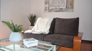 Una sala de estar con un sofá marrón, una mesa de centro de vidrio, un florero blanco con plantas verdes y una manta blanca.