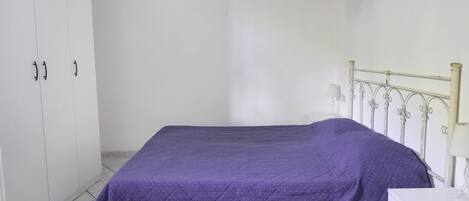A single bed with a purple bedspread, a white bedside table, and a white wardrobe with two doors.