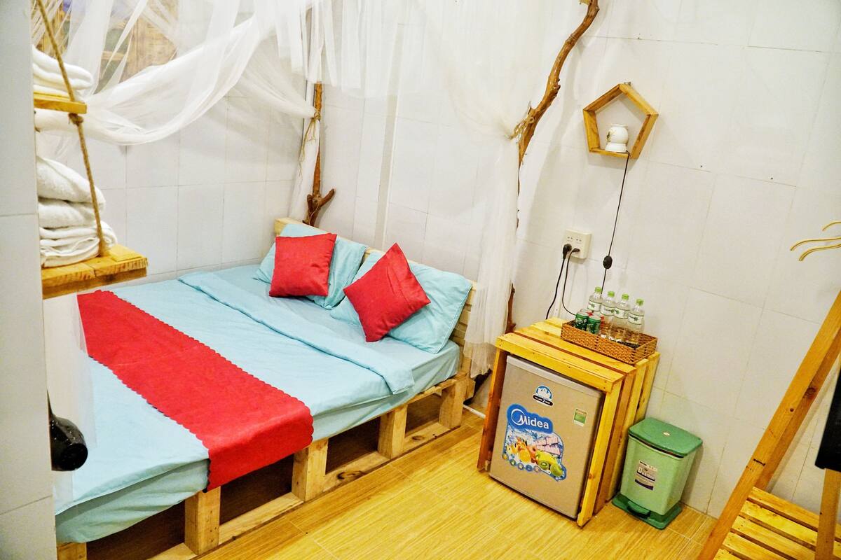 A small, rustic room with a bed, a small wooden cabinet, a trash bin, and a wall-mounted shelf.