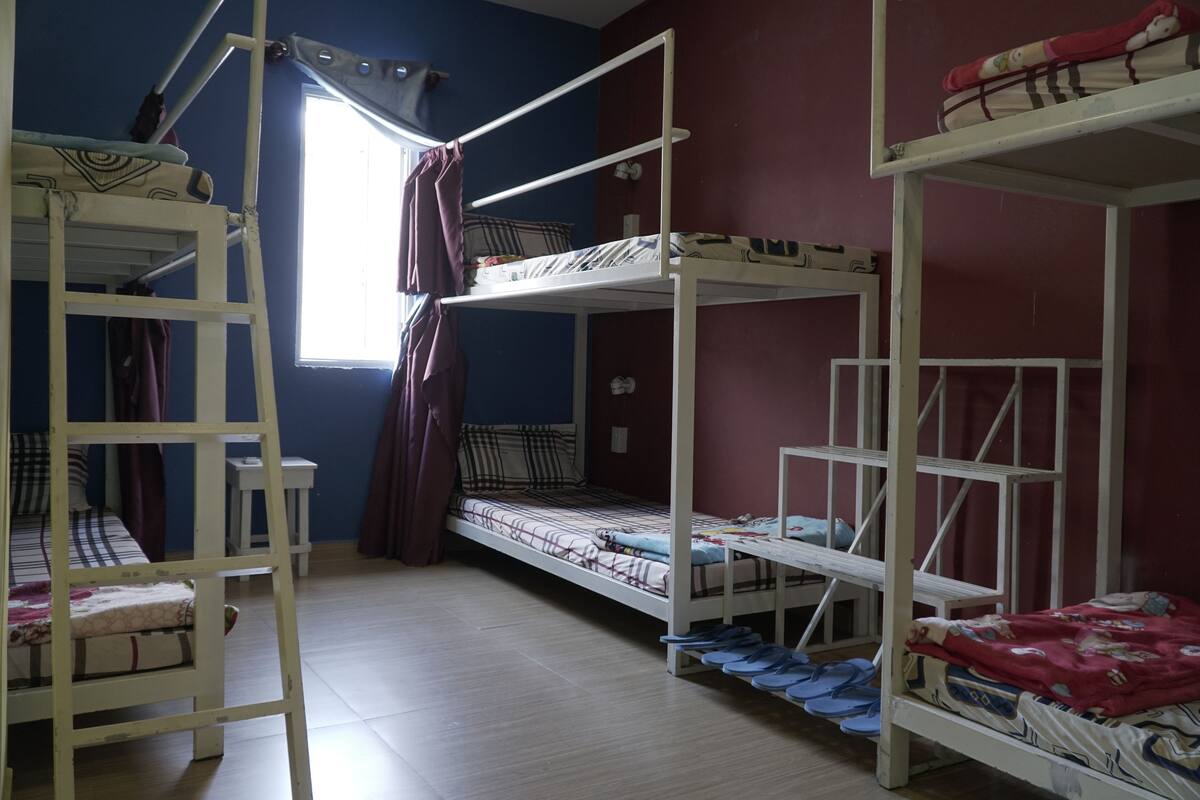 A dormitory room with bunk beds, a window with curtains, and a small table.