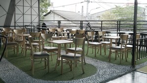 An indoor seating area with wooden chairs and tables, a green carpet, and a view of buildings and power lines outside. - Sunny Hostel (Can Tho)