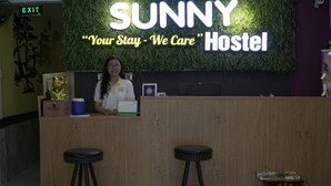 A reception desk at SUNNY Hostel with a green wall and a neon sign. There are two stools in front of the desk. - Sunny Hostel (Can Tho)