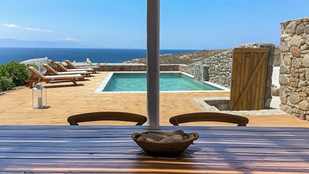 A pool area with lounge chairs and a table, overlooking a scenic view of the sea. - Emerald Bay 4BR House with pool by JJ Hospitality (Mikonos)