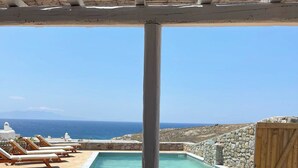A poolside area with lounge chairs, a table set for dining, and a view of the sea. - Emerald Bay 4BR House with pool by JJ Hospitality (Mikonos)