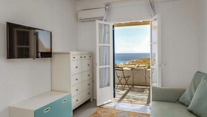A room with a TV mounted on the wall, a white dresser, a green sofa, and an open door leading to a balcony with a view of the sea. - Emerald Bay 4BR House with pool by JJ Hospitality (Mikonos)