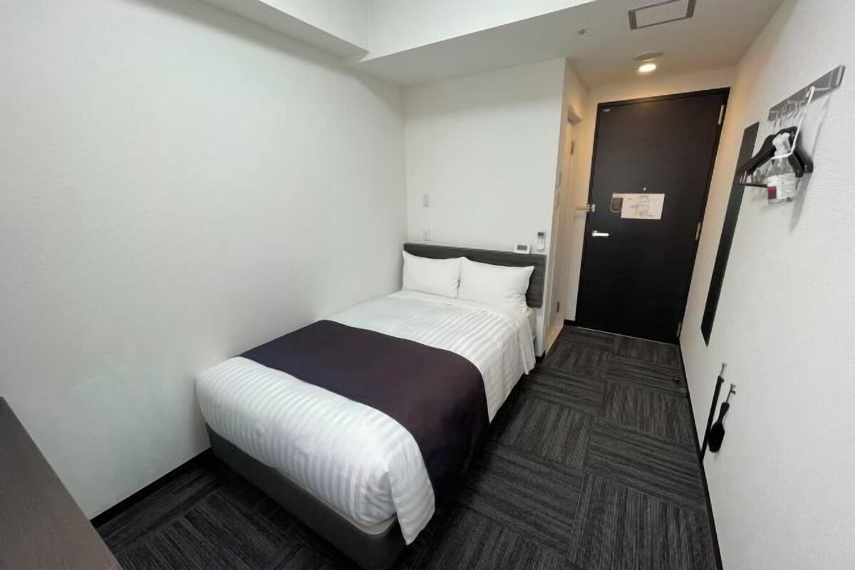 A compact hotel room with a single bed, a dark carpeted floor, and a plain white wall.