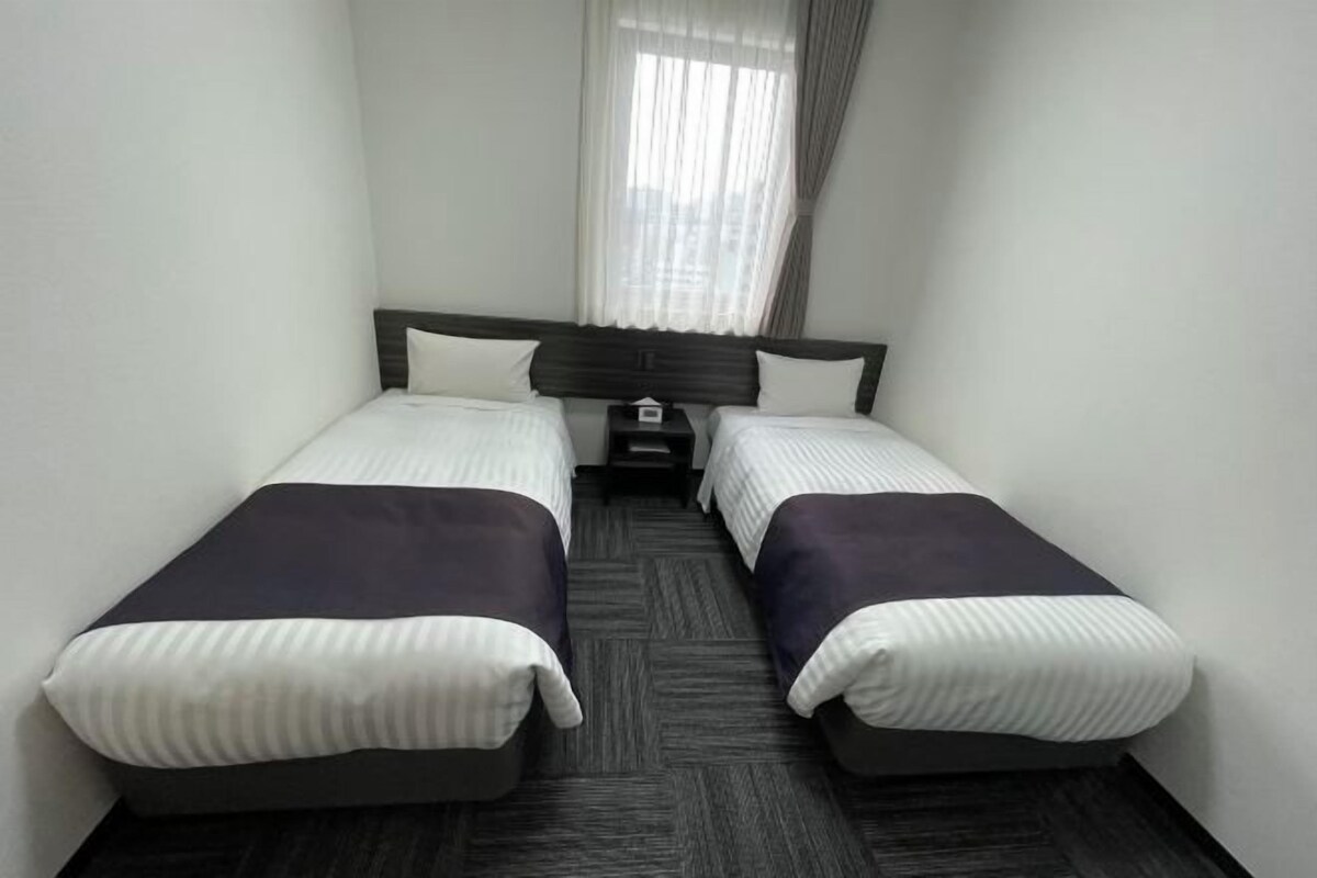 A hotel room with two single beds, a dark headboard, a small bedside table, and a window with sheer curtains.