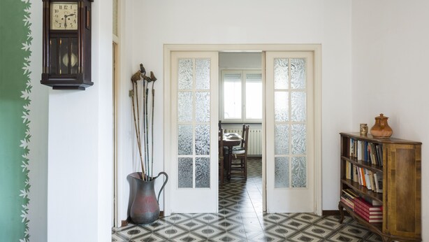 A hallway with patterned floor tiles, a wooden bookshelf, a wall-mounted clock, and a door leading to another room. - Villetta Adriana, indipendente spaziosaluminosa (Lido di Camaiore)