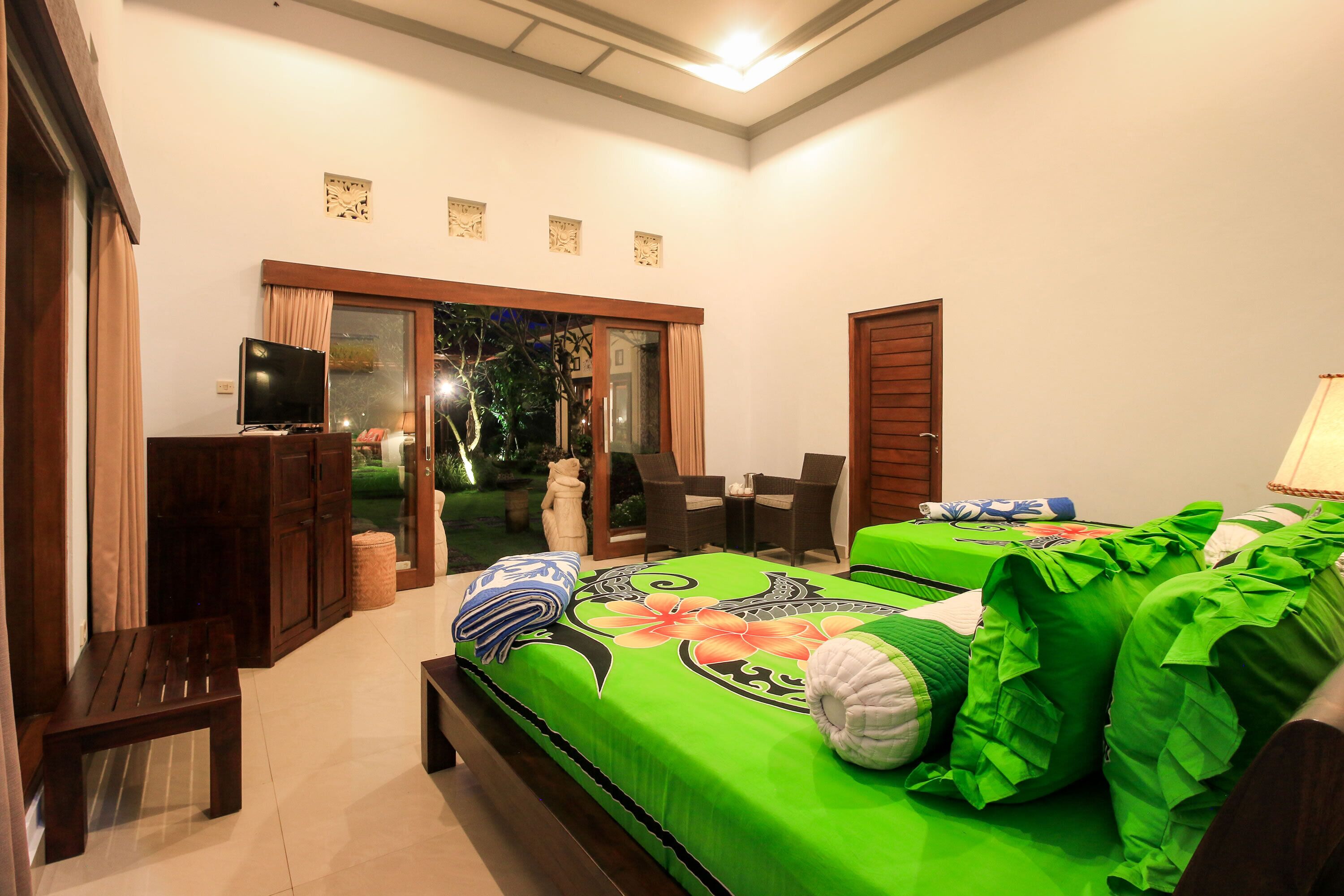 A bedroom with a green bedspread, a TV, a bench, and a view of the outdoors.