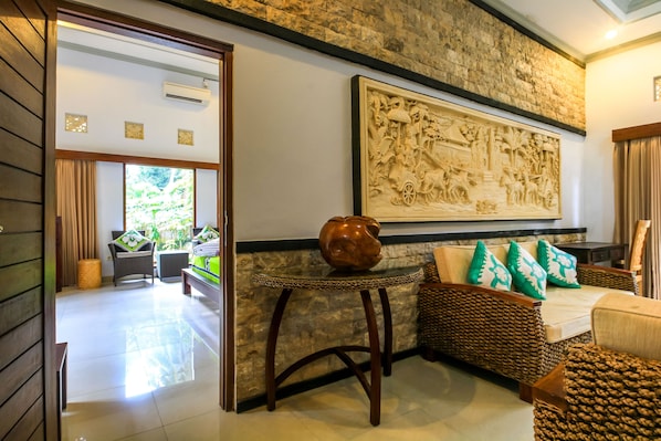 A room with a stone wall, a woven sofa with green pillows, a woven side table, and a large carved artwork above the sofa. - Asli Bali Villa (Bangli)