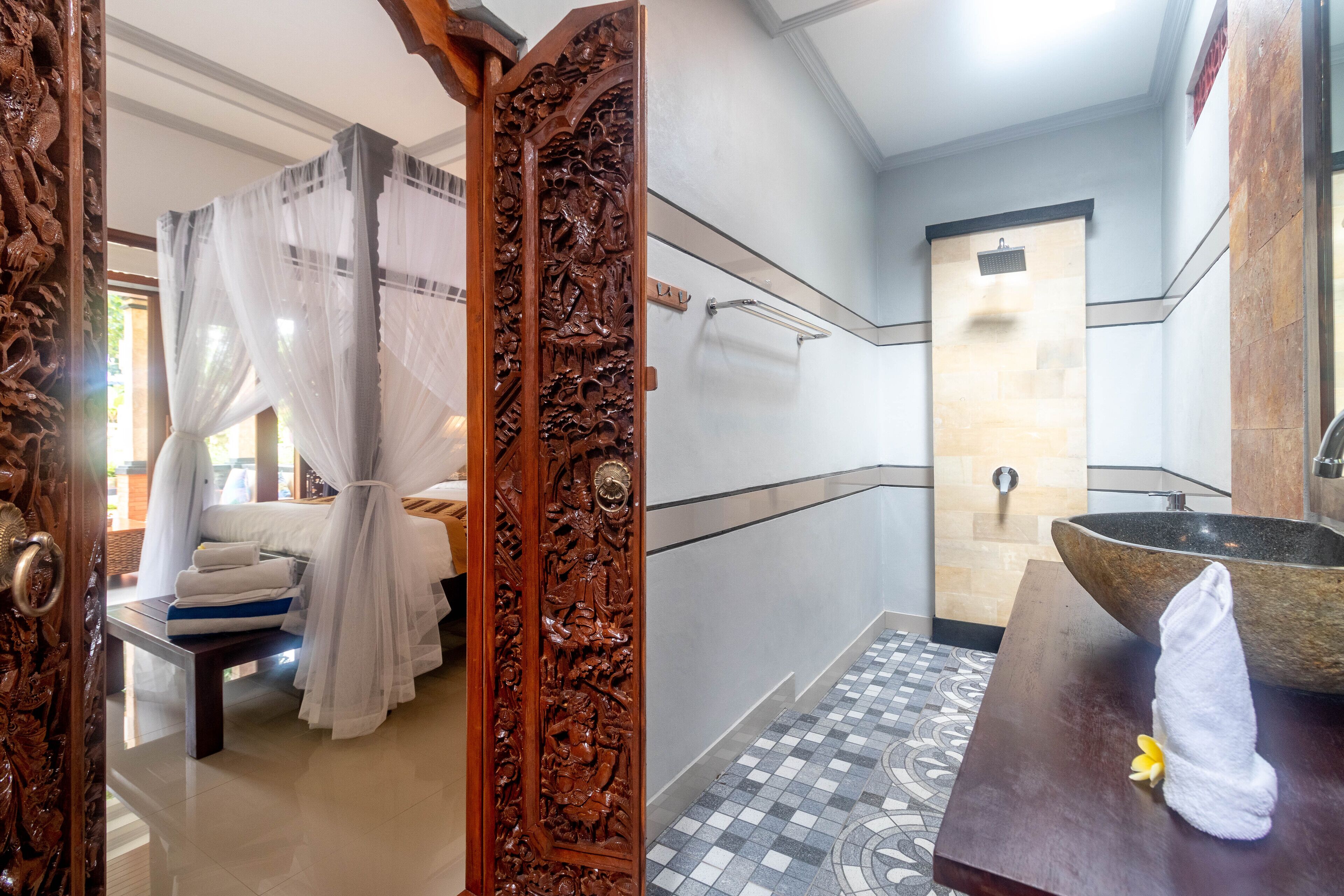 Un baño con una puerta de madera grande, una cama con sábanas blancas, un lavamanos con lavabo de piedra y un suelo de baldosas.