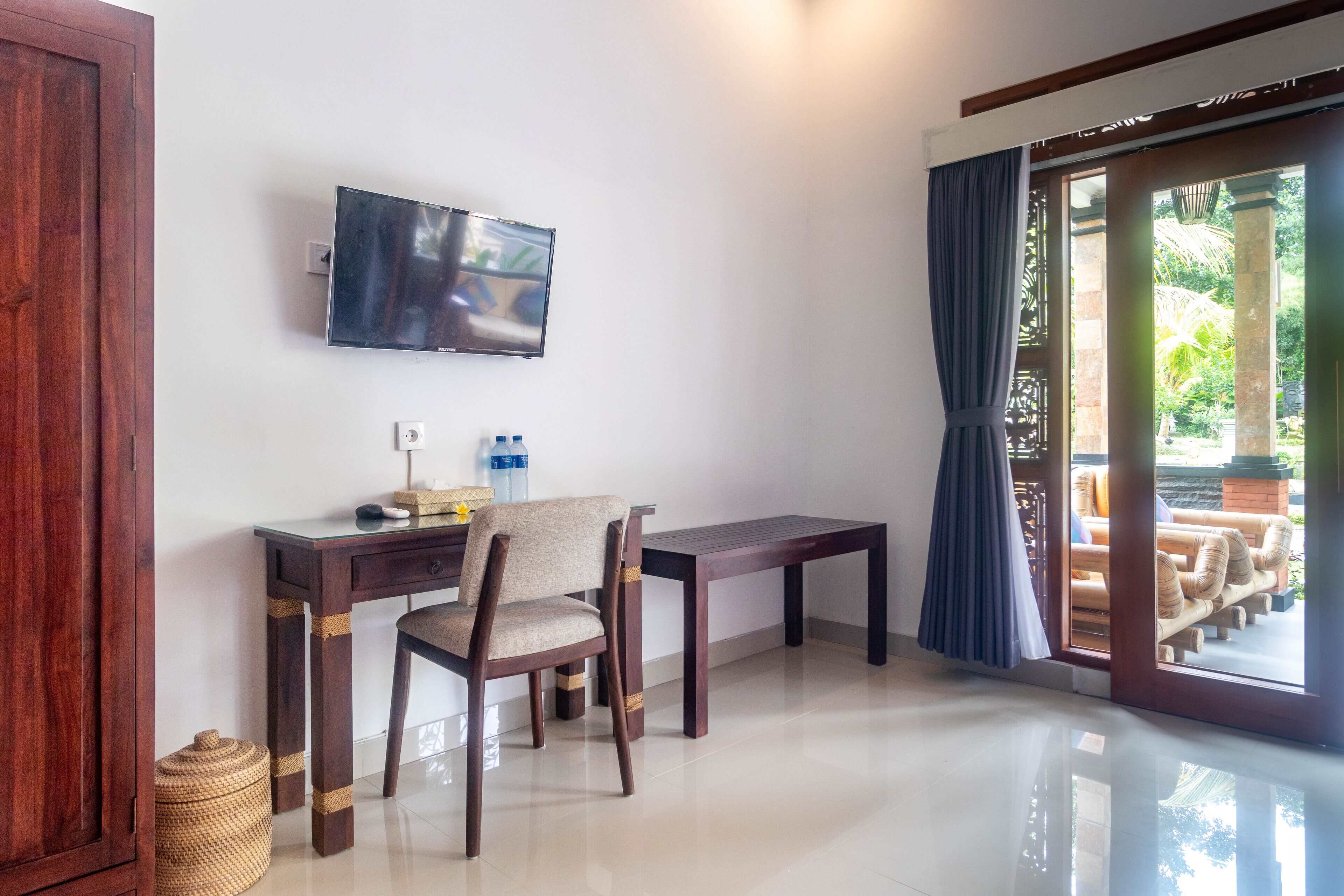 A room with a TV mounted on the wall, a wooden desk with a chair, a small table, and a view of an outdoor area through a glass door.
