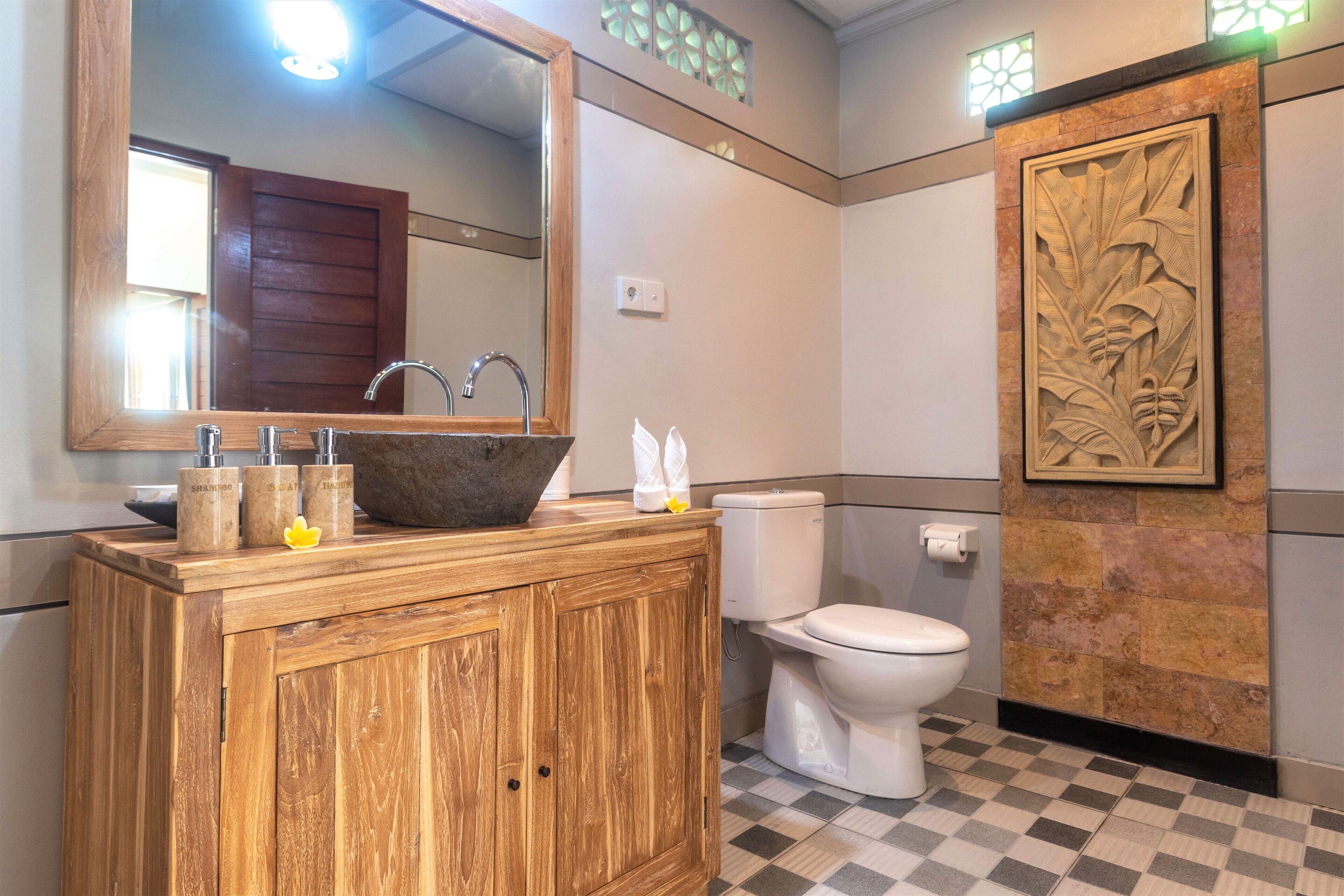 A bathroom with a wooden vanity, a large mirror, a sink with a stone basin, a toilet, and a decorative wall panel.