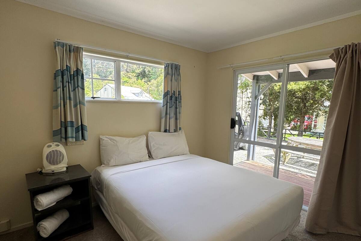 Une chambre à coucher avec un lit, une table de chevet avec un ventilateur, une fenêtre avec des rideaux et une vue sur l’extérieur.