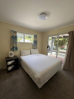 A bedroom with a bed, a nightstand with a fan, a window with curtains, and a view of the outside. - Grandeur Thermal Spa Resort (Taupo)