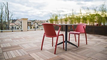 A rooftop terrace with two red chairs and a table, featuring a herringbone wooden floor and a view of buildings and trees.