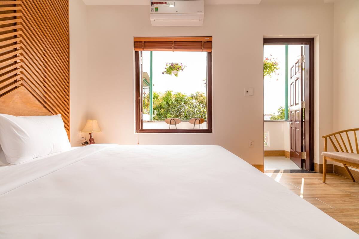 A hotel room with a large bed, a window with a view of greenery, a wooden headboard, and a bench by the door.