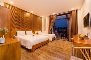 Habitación de hotel con dos camas, un cabecero de madera, un escritorio y un balcón con vistas a edificios.