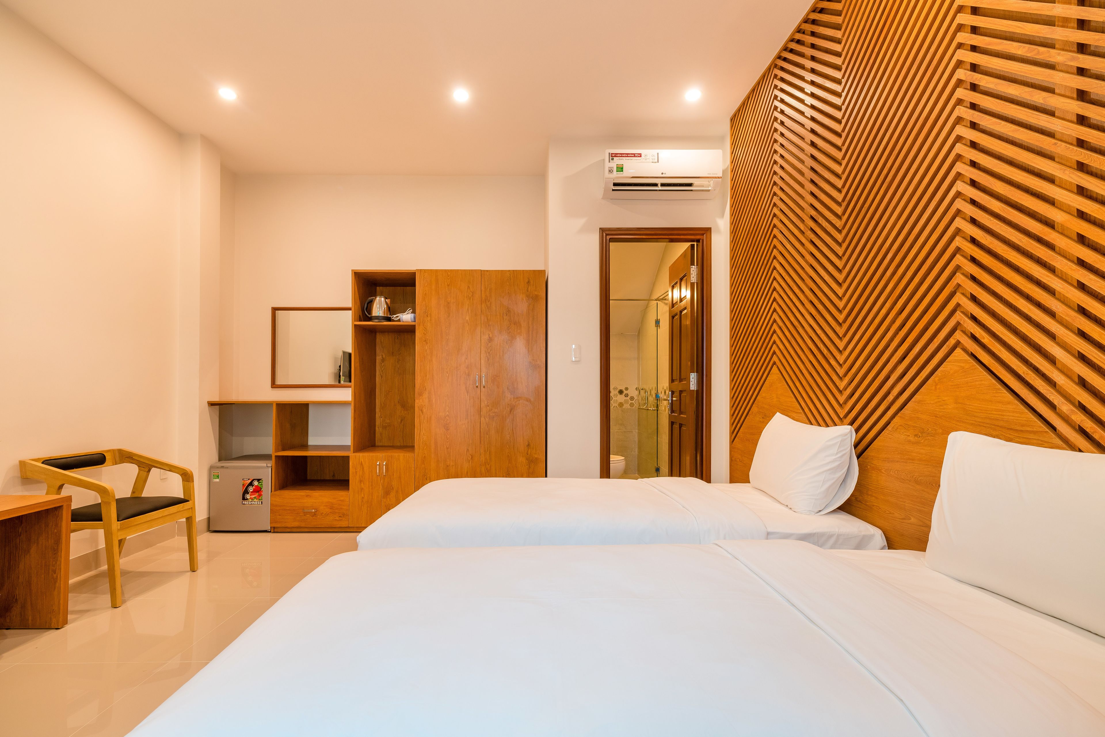 A modern hotel room with two beds, a wooden wardrobe, a desk, and a small refrigerator.