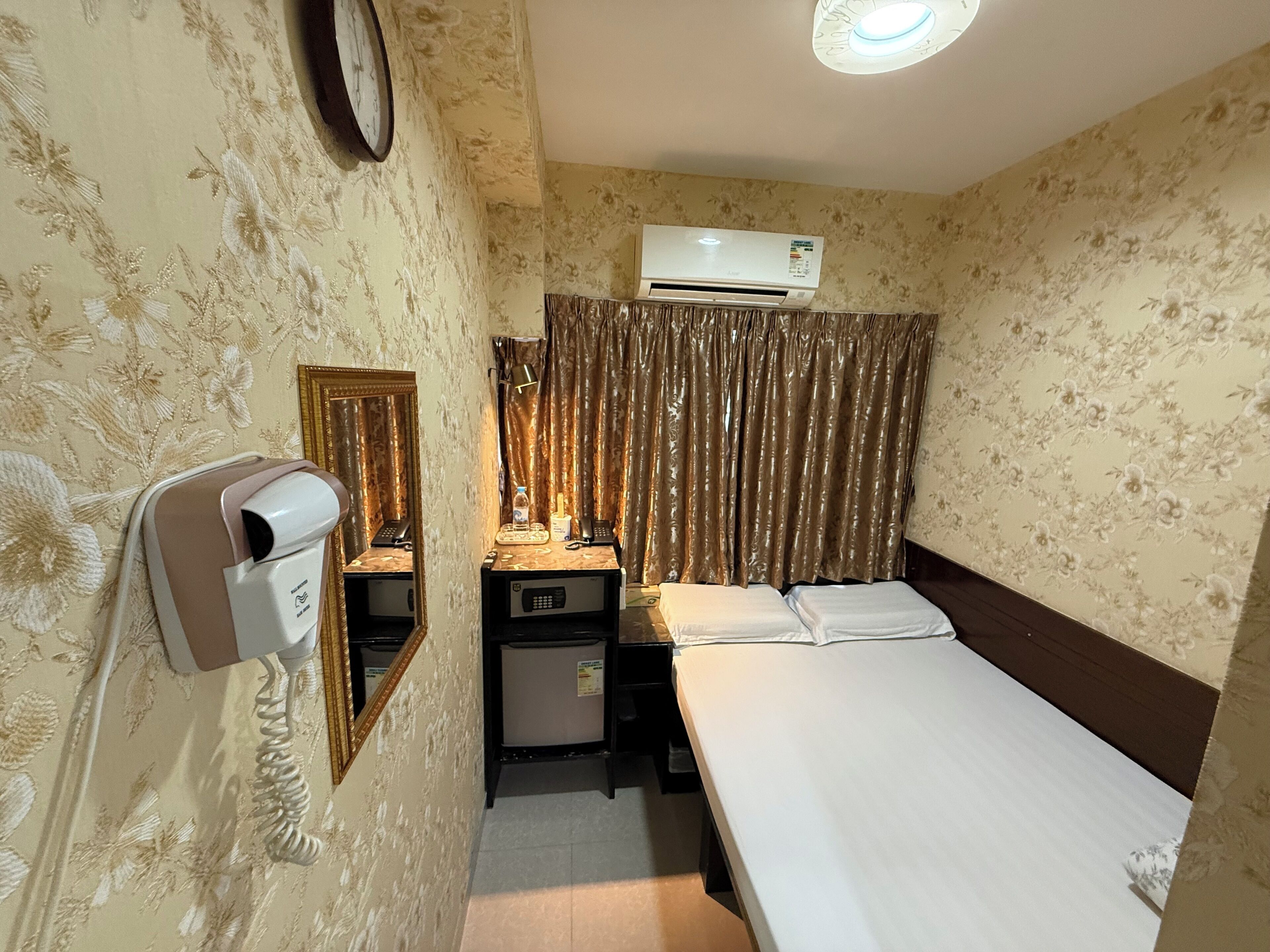 A hotel room with a bed, a desk with a telephone and a lamp, an air conditioner, and a mirror.