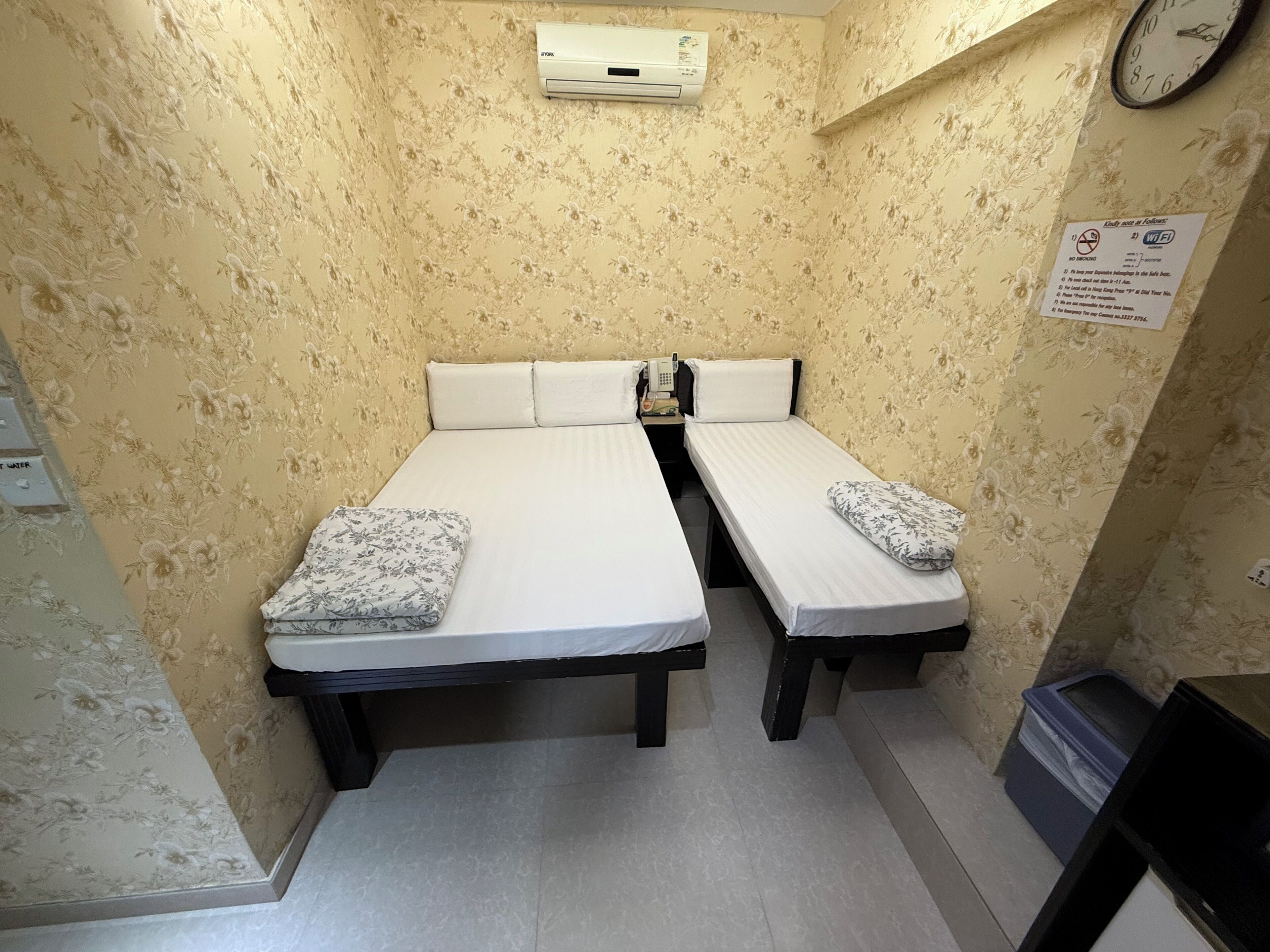 Une petite chambre avec deux lits, décorée d’un papier peint à motifs floraux, comprenant une horloge et un panneau indiquant la présence d’une connexion Wi-Fi.