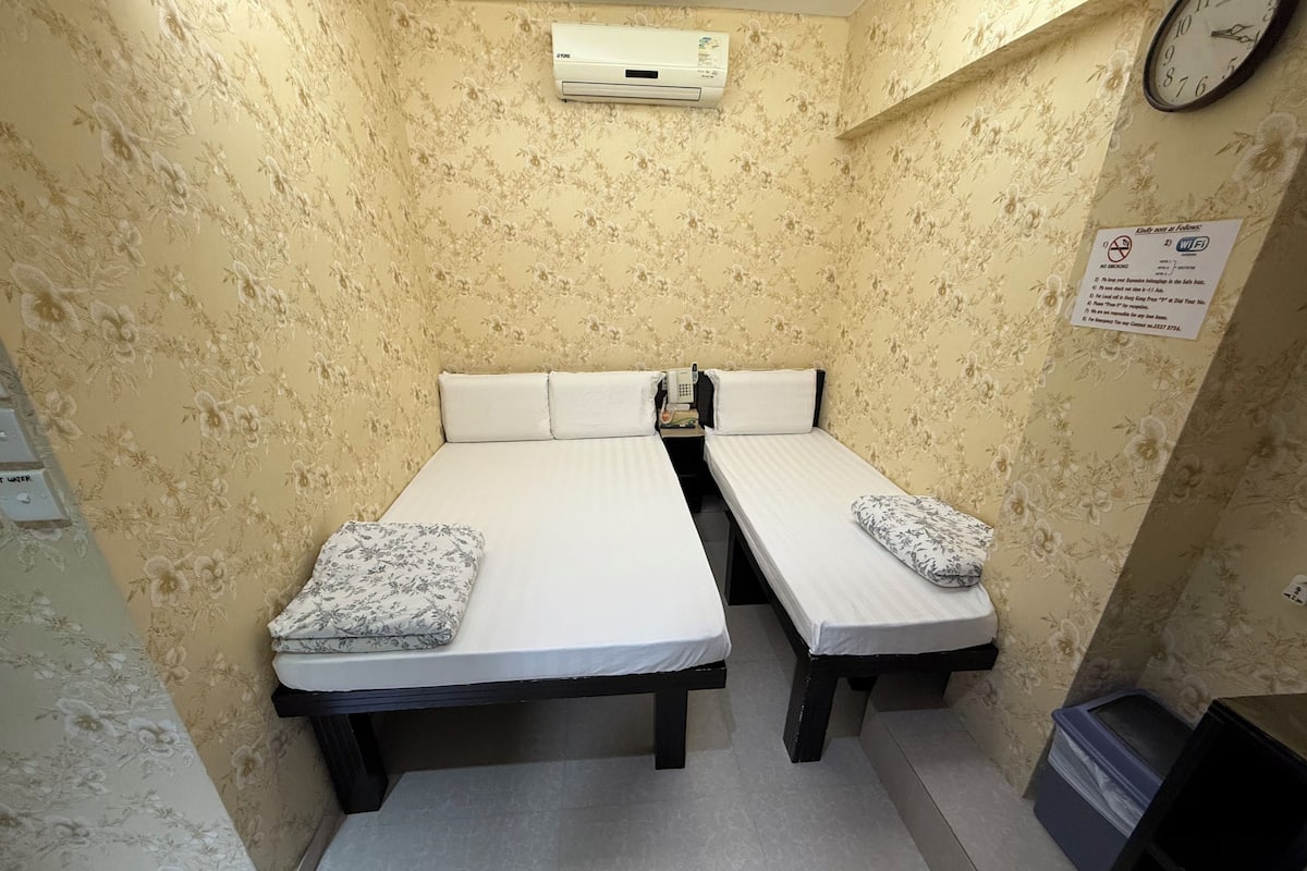 A small, two-bed room with floral wallpaper, a clock, and a sign indicating Wi-Fi availability.