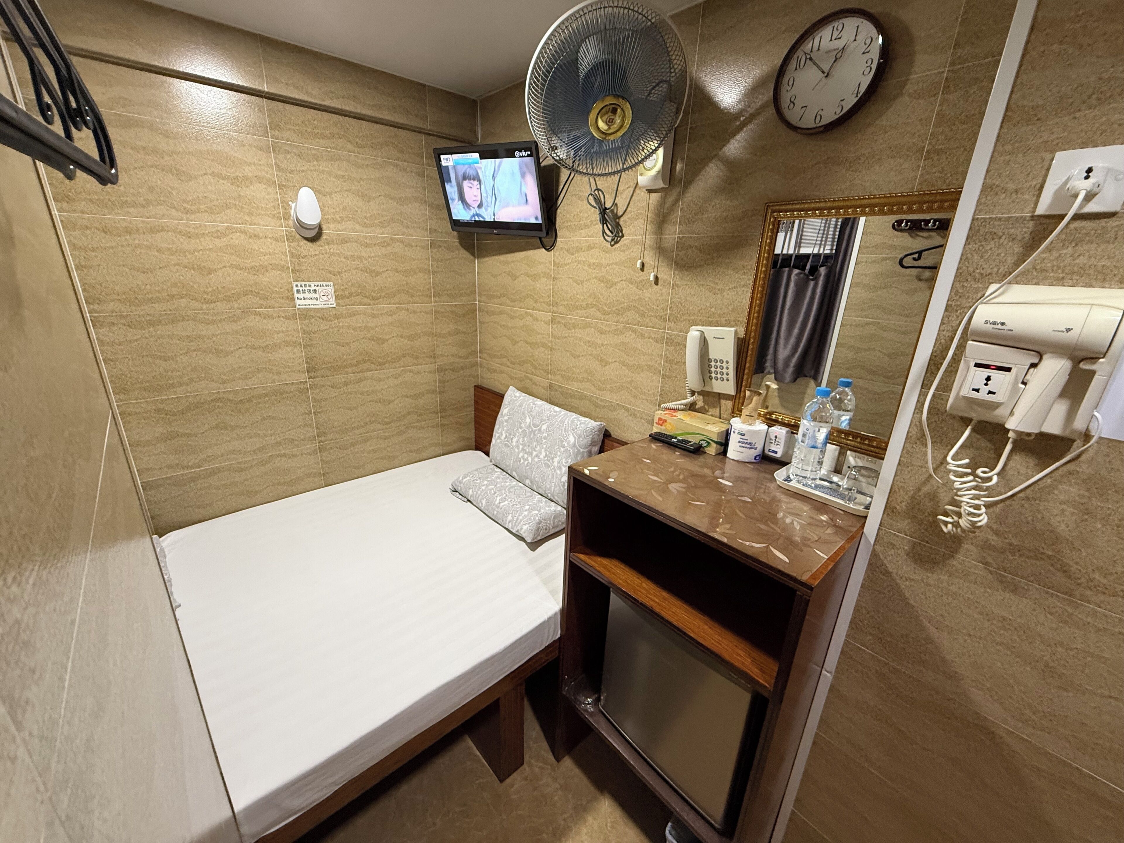 A compact hotel room with a single bed, a small bedside table with toiletries, a wall-mounted TV, a fan, a clock, and a phone.