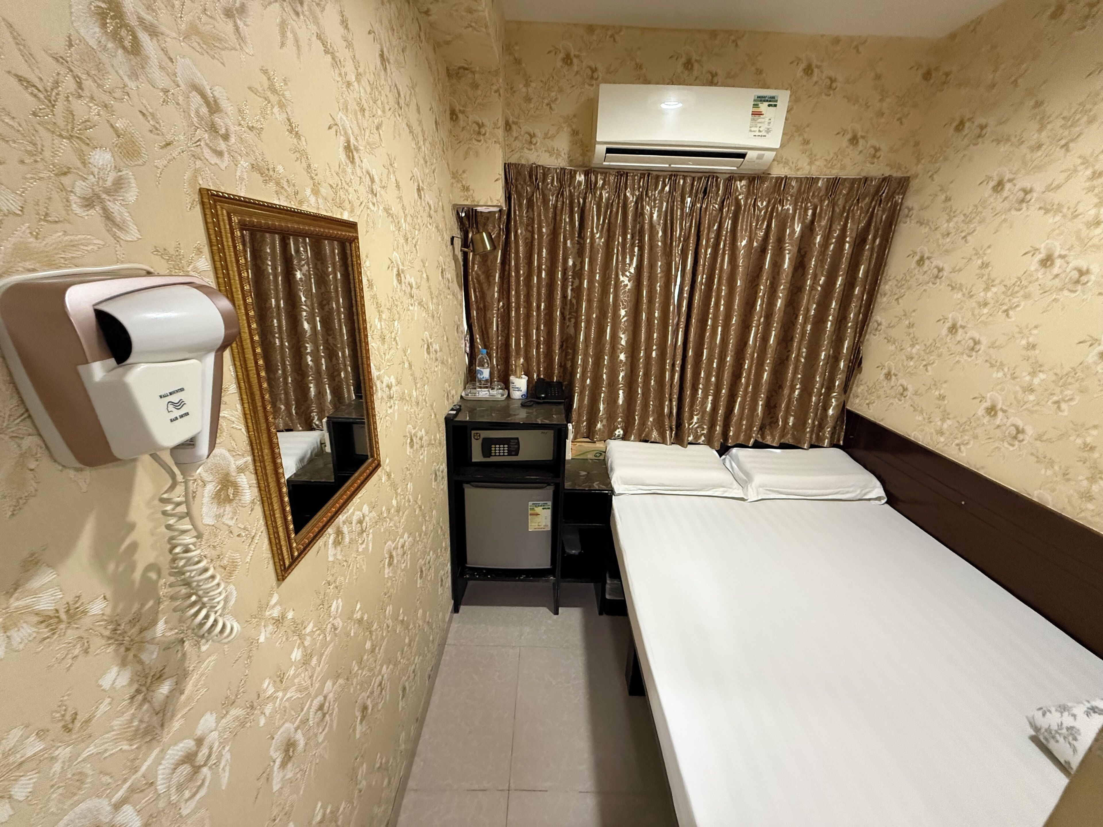 A hotel room with a bed, a desk with a mirror, a hairdryer, an air conditioner, and curtains.